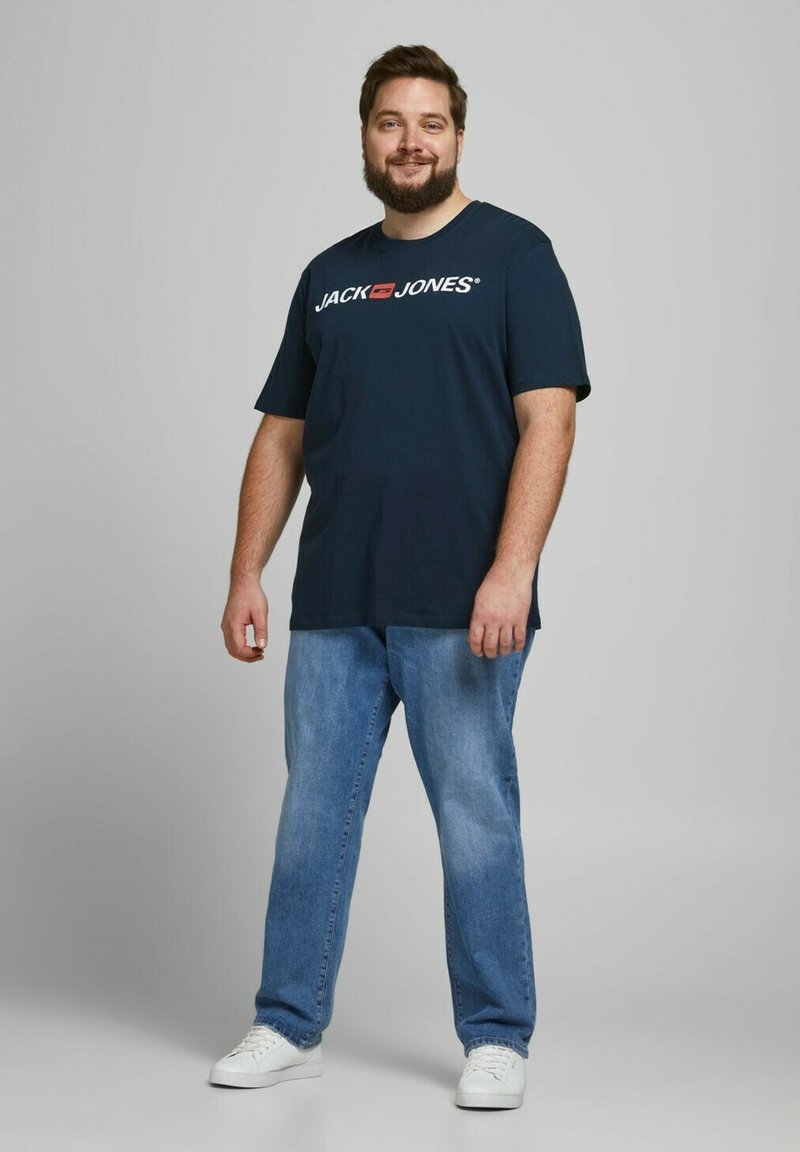 Marineblaue Baumwoll-T-Shirt mit dem "JACK & JONES"-Logo, kombiniert mit hellblauen Jeans und weißen Sneakers, vor einem grauen Hintergrund stehend.