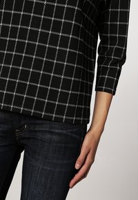 Černý top s tříčtvrtečními rukávy a bílým mřížkovým vzorem, nošený s tmavě modrými džínami, pravá ruka a ruka volně visí podél těla.