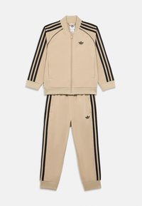 Bež trenirka s duksom na patent i suženim hlačama, obje s crnim prugama sa strane i malim logotipom na prsi i bedru.