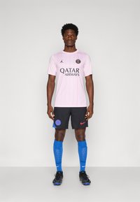 Šviesiai rožinė futbolo marškinėliai su juodais ir mėlynais akcentais, su vertikaliomis juostelėmis ir logotipu. Juodi šortai, kelių ilgio mėlyni pristatymai ir juodi sportbačiai.