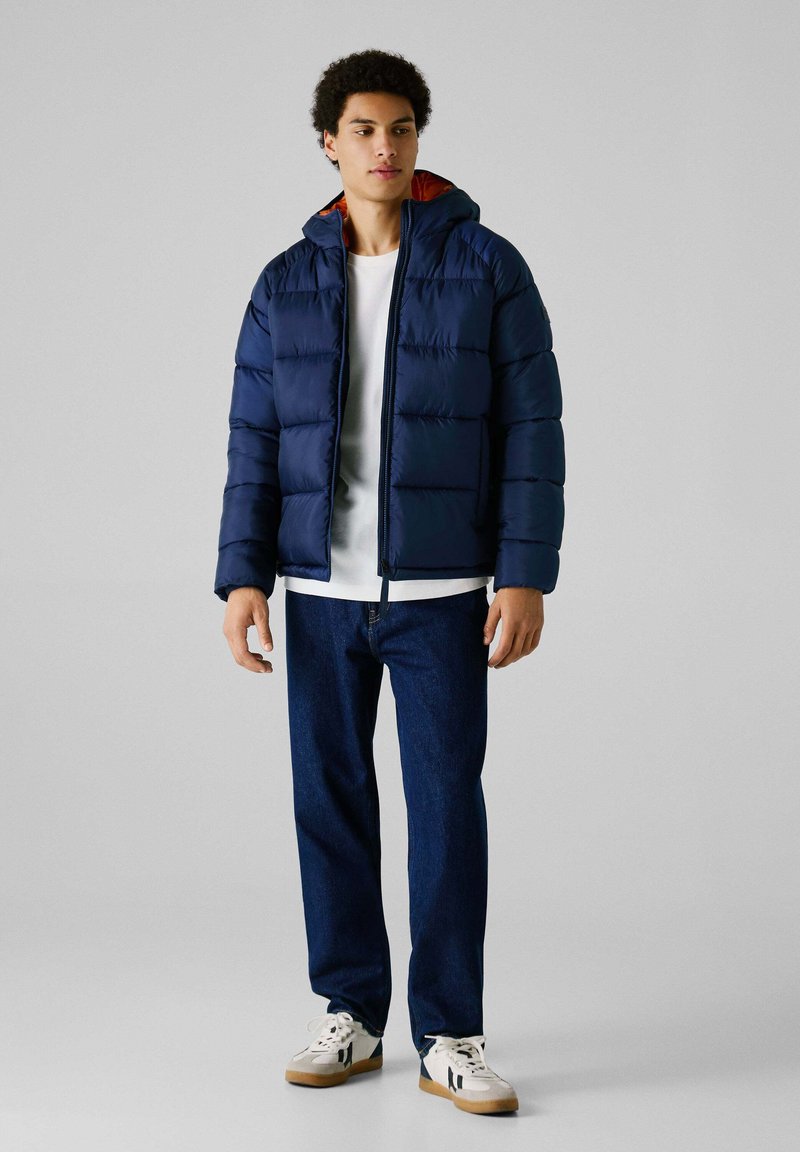 Marineblauwe pufferjack met een capuchon, voorzien van gestikte secties en een oranje voering, gecombineerd met een witte T-shirt, donkere jeans en sneakers.