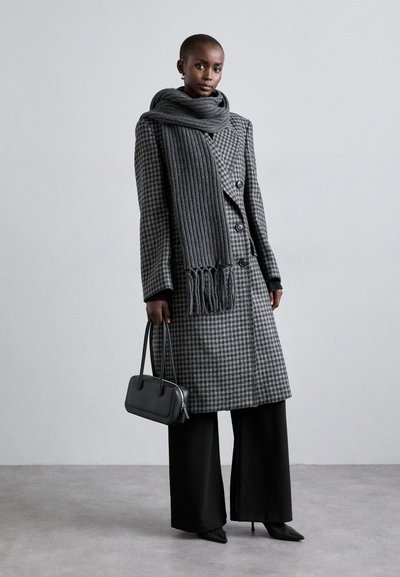 Abrigo de houndstooth gris con grandes botones, combinado con una bufanda de rayas ribeteadas en gris oscuro texturizada. Pantalones anchos negros y un pequeño bolso negro completan el conjunto.