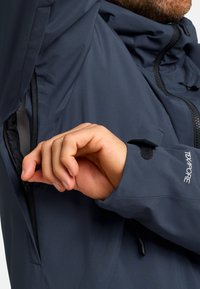 Giacca impermeabile blu navy con tasca con zip e polsini regolabili. Presenta una texture liscia e opaca e un design con colletto alto.