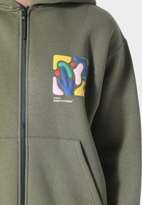 Hættetrøje med lynlås i olivengrøn fleece, med en flerfarvet abstrakt lap på brystet og to forlommer.