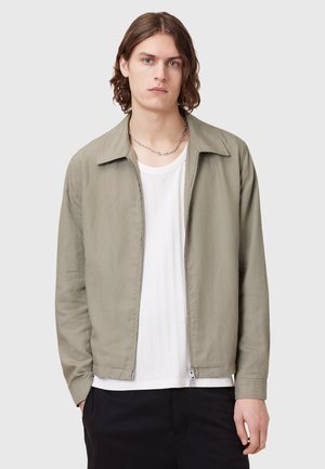 Chaqueta bomber verde claro con cremallera frontal, cuello plano y mangas largas, usada sobre una camisa blanca texturizada y pantalones negros.