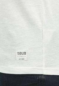 Hvit T-skjorte med teksturert stoff. Har en svart merkelapp som leser "!SOLID DENMARK EST.1999" sydd på nedre venstre side.
