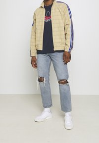 Gul rutig zip-up jacka med blå ränder, tillsammans med en marinblå t-shirt, slitna ljusblå jeans och vita sneakers.