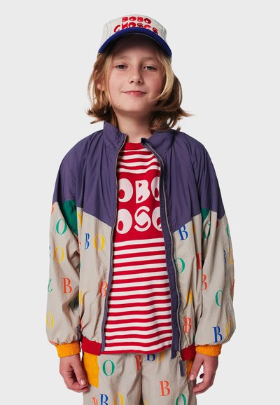 Farverig vindjakke med lilla, grønne og beige paneler, prydet med multikolor bogstaver, parret med en rød stribet t-shirt og kasket.