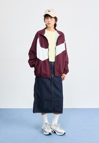 Jachetă de alergare bordeaux și albă cu mansete elastice, cămașă verde aprins, fustă din jeans, șosete albe și adidași gri deschis cu talpă neagră.
