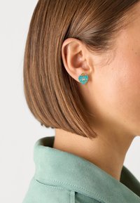 Boucles d'oreilles en forme de cœur turquoise avec des finitions dorées, arborant le texte "Guess" et un design en cœur. Portées à l'oreille droite, avec une finition lisse.