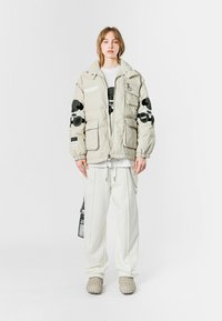 Chaqueta acolchada beige con acentos bordados en negro, que presenta múltiples bolsillos y un cuello alto. Combinada con pantalones blancos holgados y zapatos texturizados.