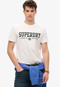 Hvid bomulds T-shirt med et stort sort "SUPERDRY" logo og "PREMIUM SPORTING" nedenunder. Akkompagneret af en blå sweater bundet om livet.