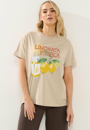 Frau trägt beige T-Shirt mit einer Grafik von Zitronen, einer Zitronenlimonade-Dose mit Strohhalm und dem Text „LIMONATA FRESCA“ und „RIPE N' READY.“