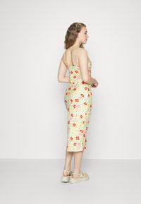 Vestido floral com alças finas, apresentando um fundo amarelo pastel e padrões florais em vermelho, rosa e verde. Silhueta ajustada até ao joelho.