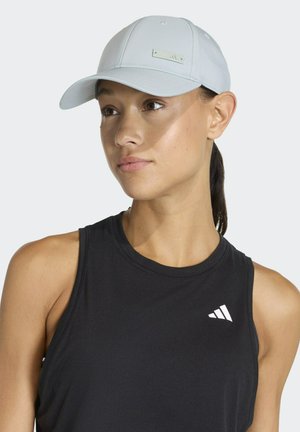 Mujer que lleva una gorra de béisbol gris claro y una camiseta sin mangas negra de Adidas, mirando ligeramente hacia un lado con una expresión neutral.