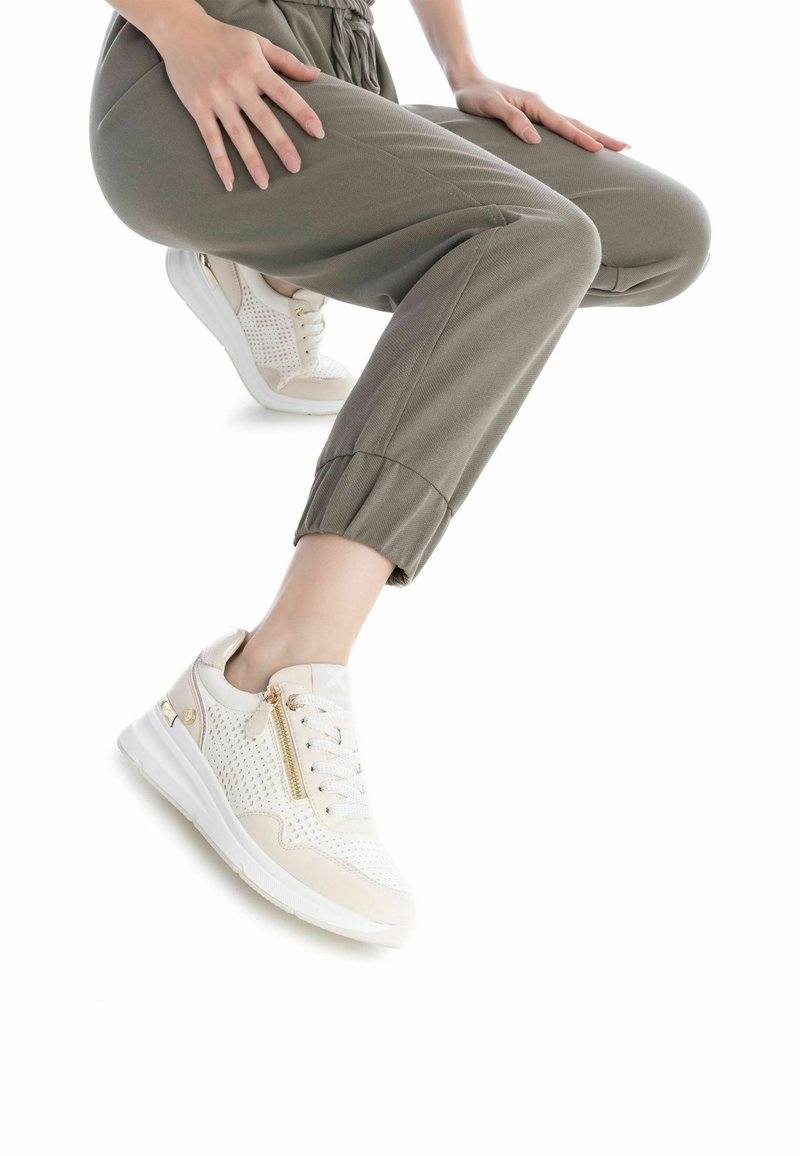 Zapatillas blancas con diseño de malla perforada, detalle de cremallera dorada y detalles de ante beige sobre una suela de goma blanca, combinadas con pantalones verde oliva.