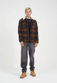 Geruite jas in donkeroranje en zwart met een witte bontkraag, gecombineerd met een grijze jeans en camelkleurige laarzen. Vier knopen en twee borstzakken.