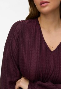 Femme à la peau claire et aux longs cheveux bruns portant un chemisier texturé à col en V de couleur violet foncé, la main reposant près de la taille.