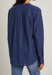 Camicia a maniche lunghe blu navy realizzata in tessuto leggero, con una vestibilità ampia, plissettatura sul retro e polsini con bottone. Superficie liscia e testurizzata.