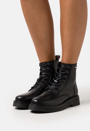 Černé kožené kotníkové boty s kulatou špičkou, plochou podrážkou, provlečeným šněrováním a malým logem na boku. Hladká textura a robustní design.