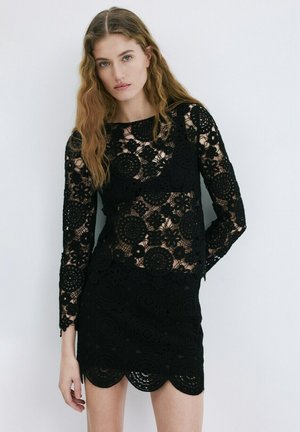 Mujer con cabello largo y ondulado, vestida con una blusa de encaje negra de manga larga y una falda a juego de encaje negro, sobre un fondo blanco.