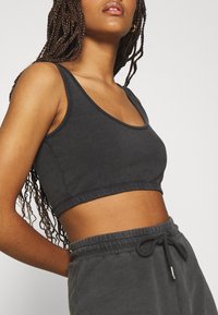 Femme portant un débardeur court noir et un pantalon à cordon assorti, avec de longs cheveux tressés sur un fond uni.