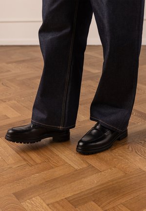 Person, die dunkelblaue Jeans mit weitem Bein und schwarze Lederloafers trägt und auf einem Holzdielenboden steht.