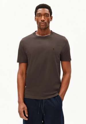 Homme portant un t-shirt marron foncé à manches courtes et un pantalon bleu foncé, debout avec une main dans la poche sur un fond uni.