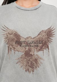 Grijze katoenen t-shirt met een grote bruine arendgrafiek en tekst in zwart die luidt: "JUST CAVALLI FIRED EARTH".