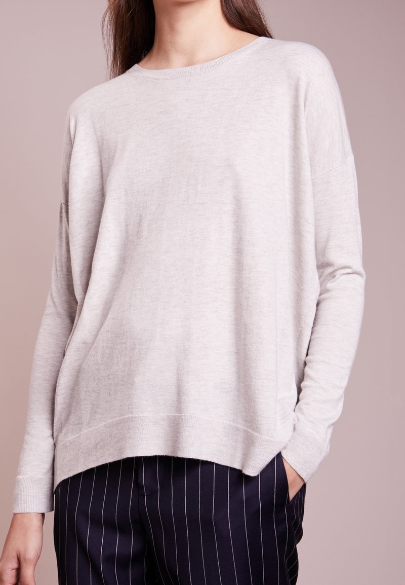 Personne portant un pull beige clair à manches longues avec une coupe décontractée et un pantalon foncé à fines rayures, debout avec une main dans la poche devant un fond uni.