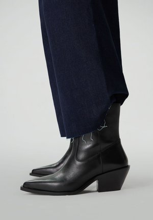 Bottines en cuir noir avec un bout pointu et un talon épais, complétées par un pantalon en denim foncé avec un ourlet effiloché.