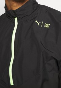 Svart vindjacka med hög krage, med en grön dragkedja och logotyp. Slät textur, lätt material och subtila paneldesigndetaljer.