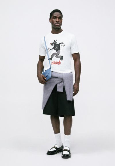 Balta T-krekls ar vilka grafisko attēlu, saskaņots ar melnām šortām, gaiši pelēku džemperi, kas saistīts ap vidukli, un zilu pleca somu. Melni loaferi ar baltām zeķēm.