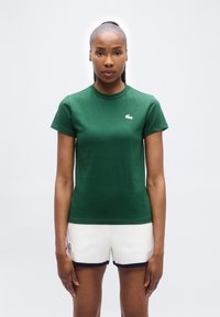 Γυναίκα που φοράει πράσινο πουκάμισο με κοντά μανίκια και λευκά σορτς με μαύρο περίγραμμα, στέκεται μπροστά σε απλό φωτεινό φόντο, κοιτώντας μπροστά.