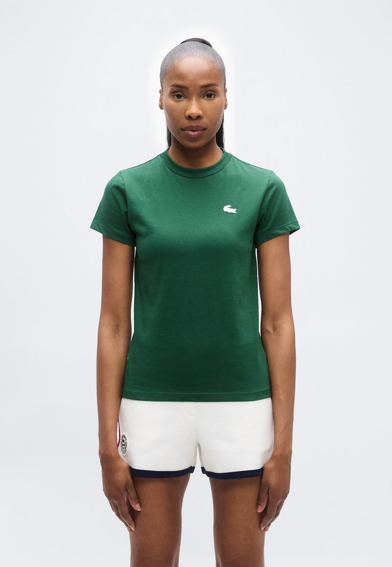 Γυναίκα που φοράει πράσινο πουκάμισο με κοντά μανίκια και λευκά σορτς με μαύρο περίγραμμα, στέκεται μπροστά σε απλό φωτεινό φόντο, κοιτώντας μπροστά.