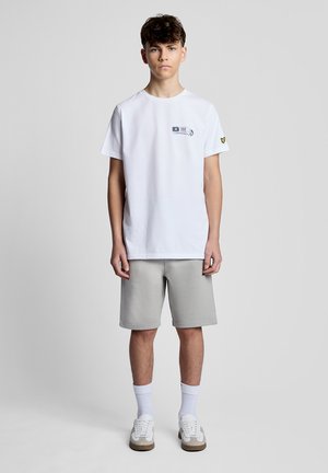 Tienerjongen staat recht voor zich uit kijkend, gekleed in een wit T-shirt met print, grijze korte broek, witte sokken en witte sneakers, tegen een effen achtergrond.
