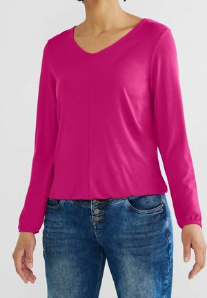 Vrouw draagt een felroze blouse met lange mouwen en een V-hals, gecombineerd met blauwe spijkerbroek met drie knopen en zakken.