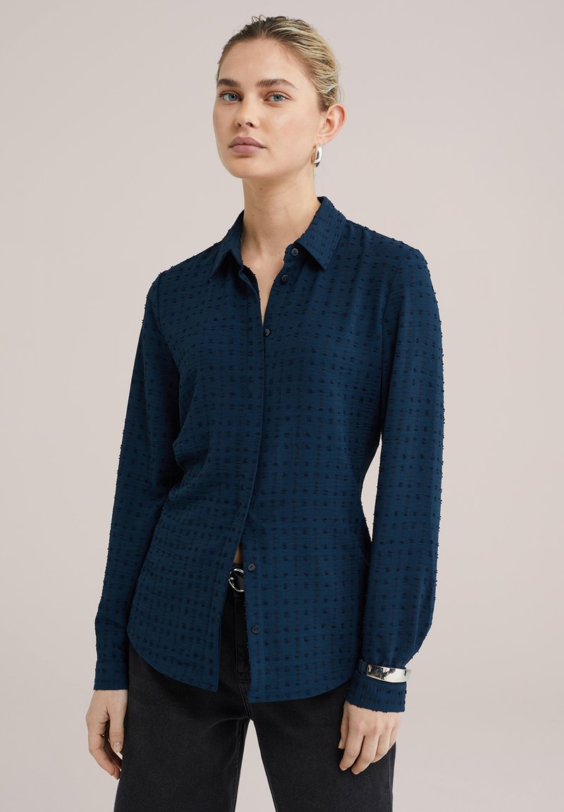 Camisa de botones azul marino con un patrón texturizado, mangas largas, cuello puntiagudo y un ajuste relajado. Combinada con unos pantalones vaqueros negros.