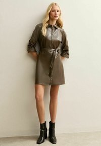 Vrouw draagt een olijfgroene nepleren overhemdjurk met ceintuur en zwarte enkellaarsjes, staand tegen een effen beige muur.