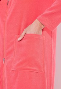 Prenda de forro polar con cremallera y un bolsillo. La tela es suave y texturada, con una cremallera oculta y mangas largas.