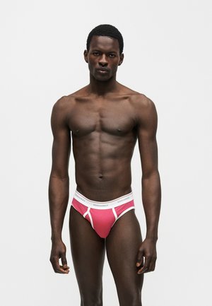 Γυμνασμένος σκουρόχρωμος άνδρας στέκεται μπροστά σε απλό φόντο φορώντας ροζ και λευκά εσώρουχα DSQUARED2, κοιτάζοντας ευθεία με ουδέτερη έκφραση.