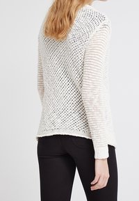 Pull léger en tricot crème avec un motif en maille texturée et aérée. Présente des manches longues et un ourlet arrière légèrement plus long.