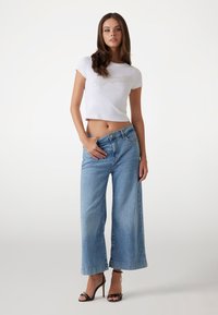 T-shirt bianco cropped con logo "Guess", jeans in denim azzurro a gamba larga e sandali neri con tacco alto. Tessuto morbido e vestibilità rilassata.