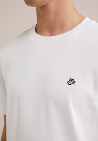 Moški nosi enobarvno belo majico s majhnim črno-belih ptičkom, izvezenim na levem prsnem delu, na nevtralni podlagi.