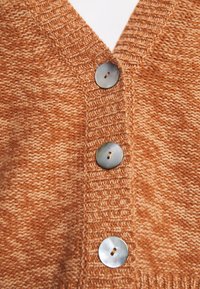 Detailní záběr oranžové pletené látky se třemi kulatými, lesklými knoflíky se dvěma dírkami, které jsou svisle připevněné podél žebrovaného okraje.