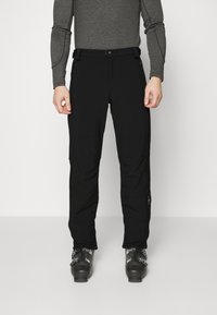 Black ski pants made of flexible fabric, featuring a slim fit, zip pockets, and reinforced hems, paired with black ski boots.