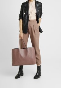 Suuri laventelinvärinen nahkainen olkalaukku, jossa on teksturoitu pinta, kaksinkertaiset kahvat ja jämäkkä muoto. Yhdistetty mustaan nahkaiseen bleiseriin ja ruudullisiin housuihin.