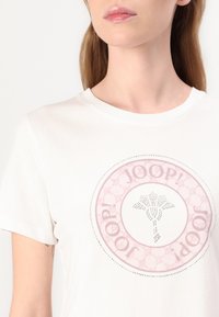 Balta medvilninė marškinė su apvaliu rožiniu logotipo dizainu, kuriame yra tekstas "JOOP!" ir centrine gėlių grafika, apjuosta cirkoniais.