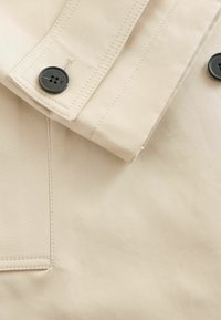Beige stofjakke med foldet krave, sort knap og synlig syning, med en glat tekstur og et minimalistisk design.