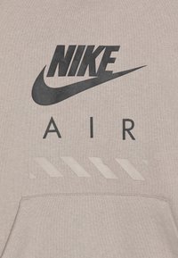 Sudadera gris claro con un gran logo negro de "Nike" y el texto "AIR". Presenta una franja inferior con acentos diagonales en blanco. Textura suave.